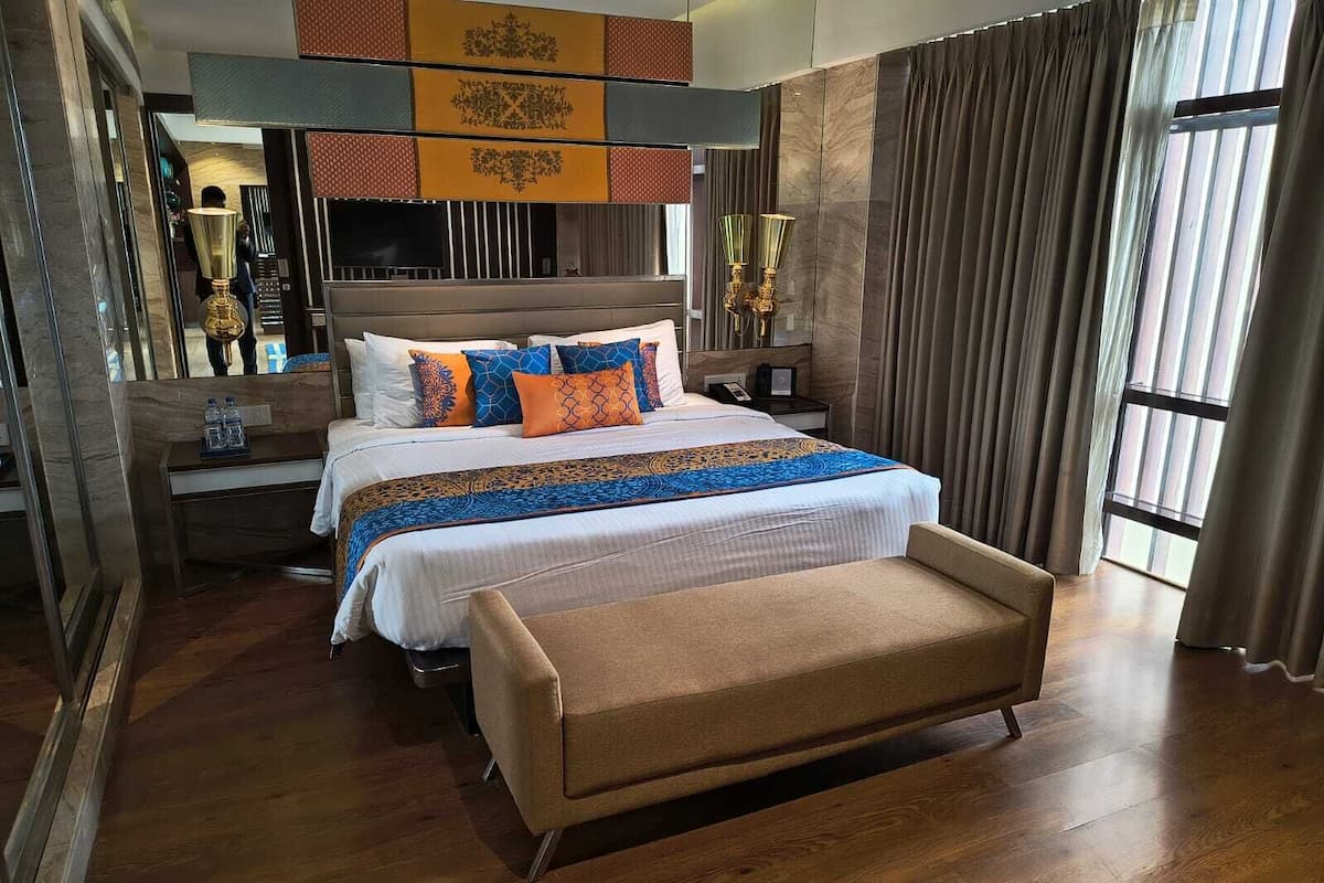 Quarto de hotel com uma cama grande, uma mesa de cabeceira, um banco e uma janela ampla com cortinas.