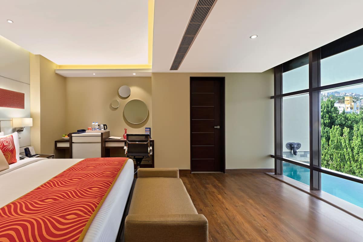 Quarto de hotel moderno com uma cama grande, uma escrivaninha, uma cadeira e vista para a cidade através de amplas janelas.