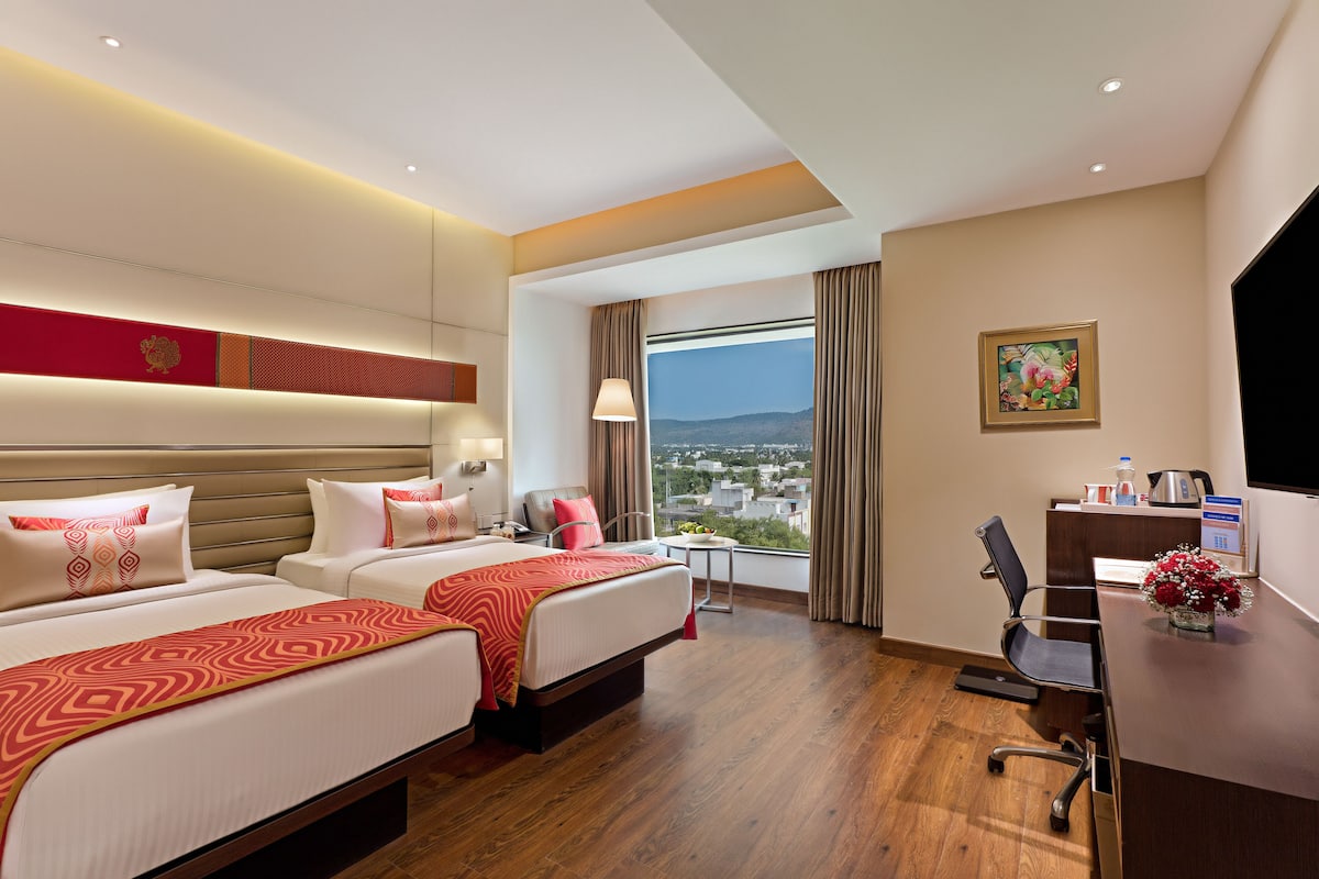 Quarto de hotel com duas camas, uma escrivaninha, uma cadeira, uma mesinha, janela com vista e um quadro na parede.