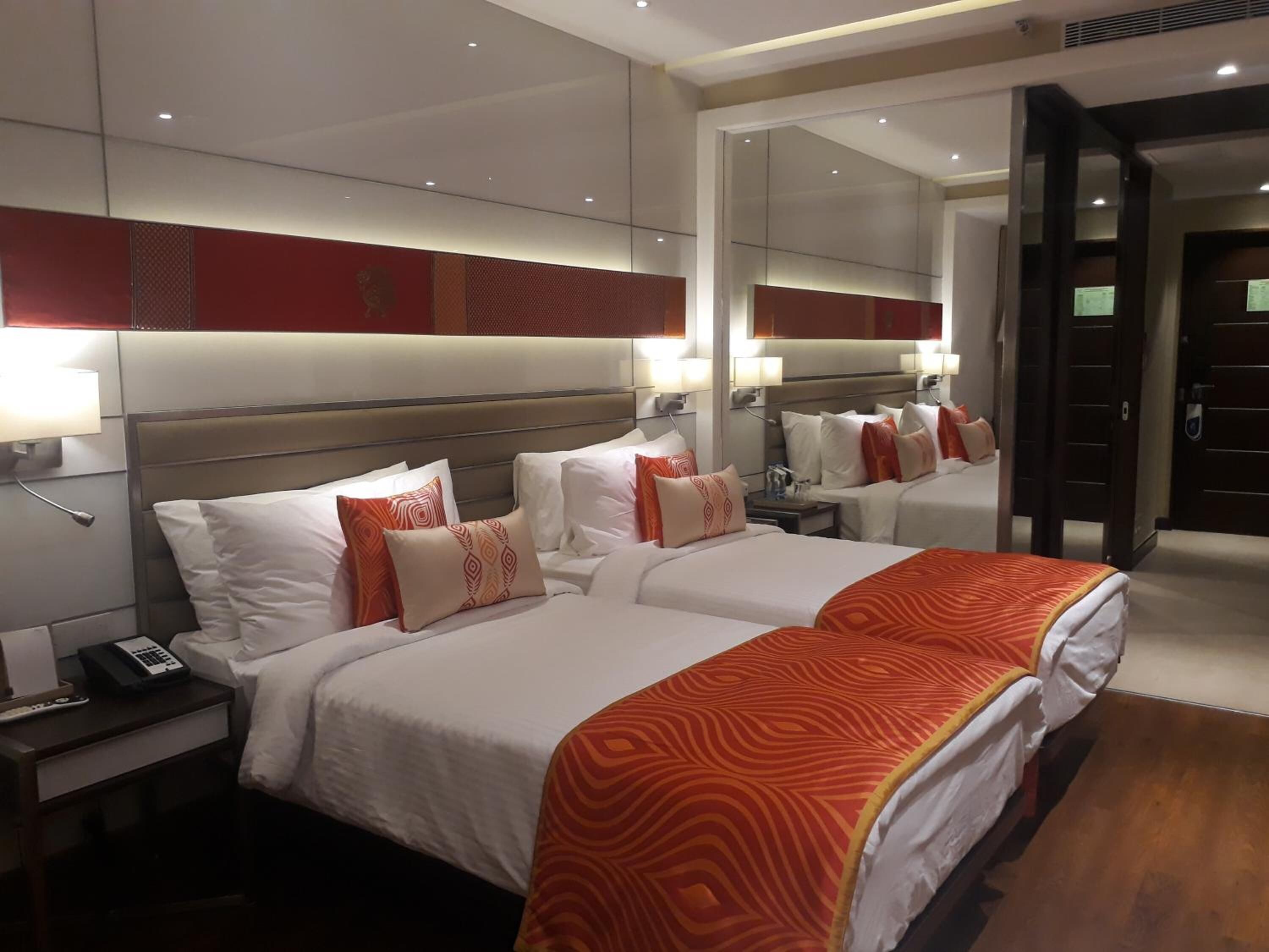 Habitación de hotel con dos camas, cada una con sábanas blancas y mantas anaranjadas estampadas. Hay una mesita de noche con un teléfono y una lámpara. La habitación tiene un espejo grande y una puerta que conduce a otra habitación.