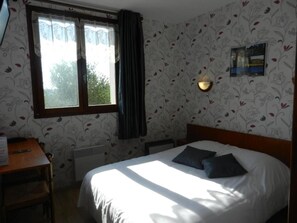 Ein Schlafzimmer mit Bett, Schreibtisch, einem Fenster mit Vorhängen und einer Wandleuchte.