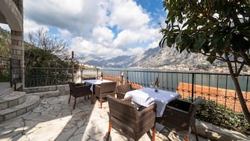 A terrace with wicker furniture and a table set for two, overlooking a lake and mountains.