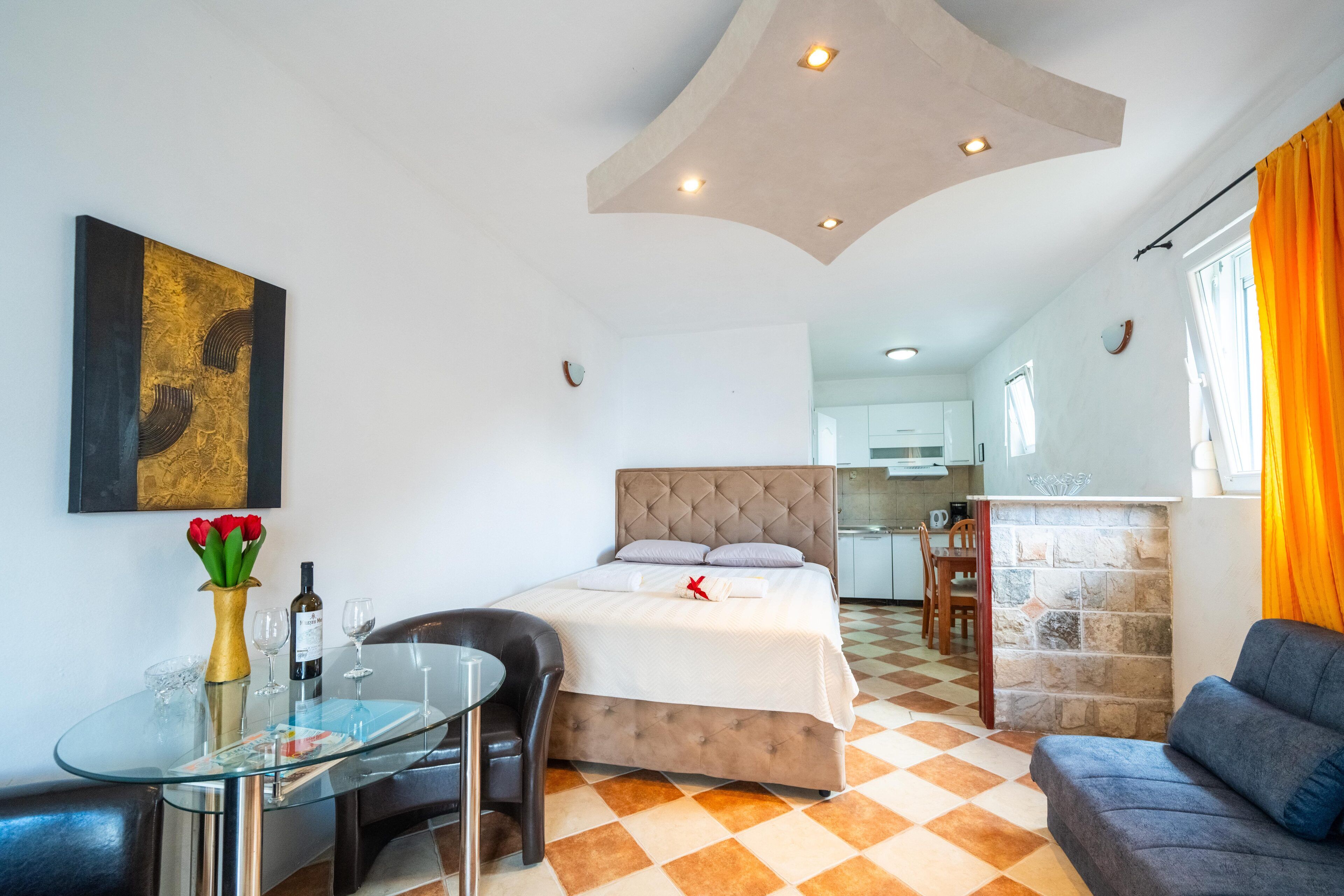 A modern hotel room with a bed, a glass dining table, a sofa, and a kitchen area in the background.