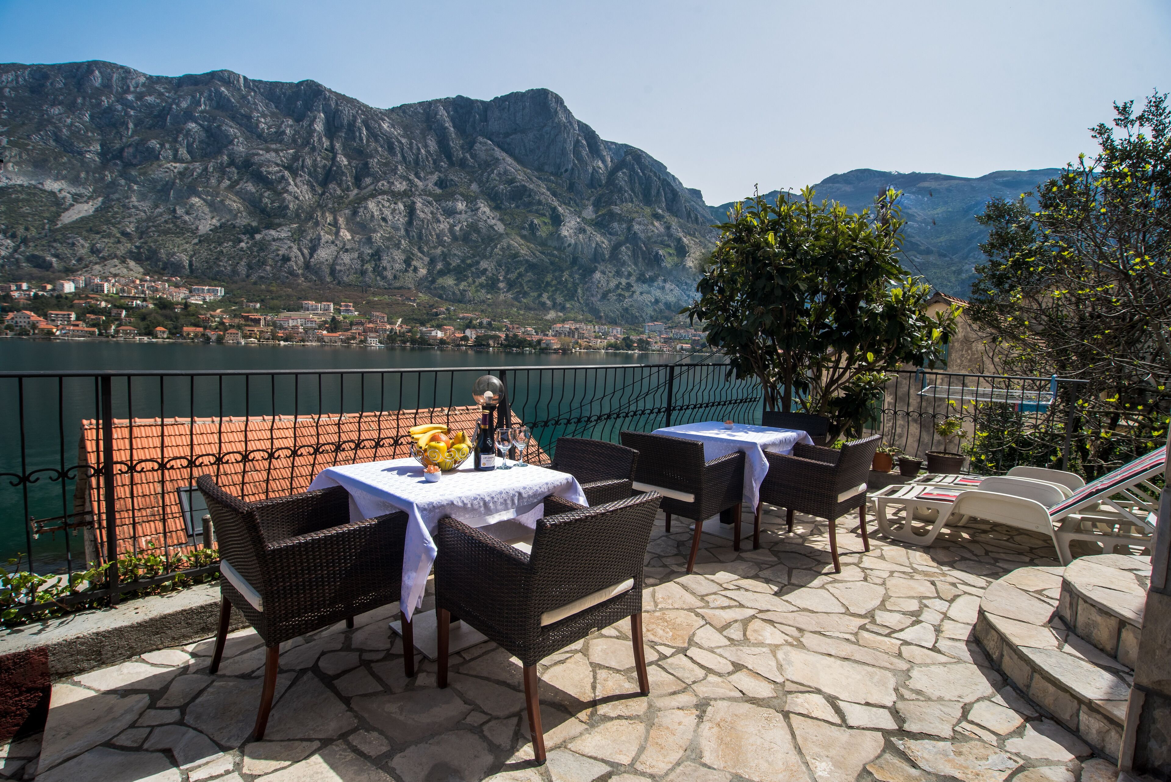 A terrace with wicker furniture and tables, overlooking a lake and mountains.