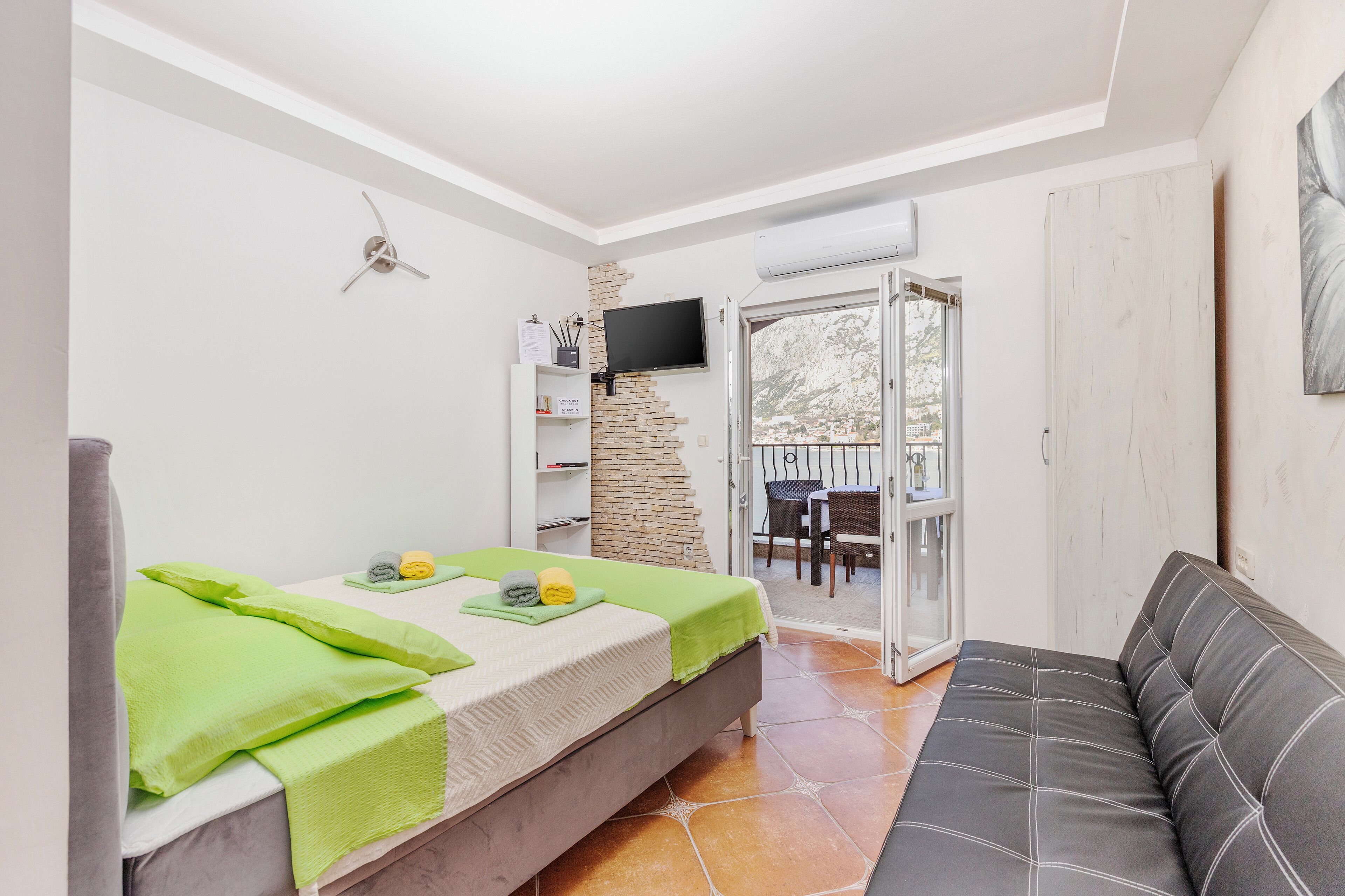 Une chambre à coucher avec un lit, un canapé gris, une télévision et un balcon équipé d’une table et de chaises.