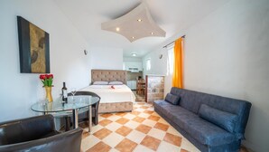 Habitación de hotel con cama, sofá, mesa de vidrio con sillas, pequeña mesa de comedor y un jarrón decorativo con flores.