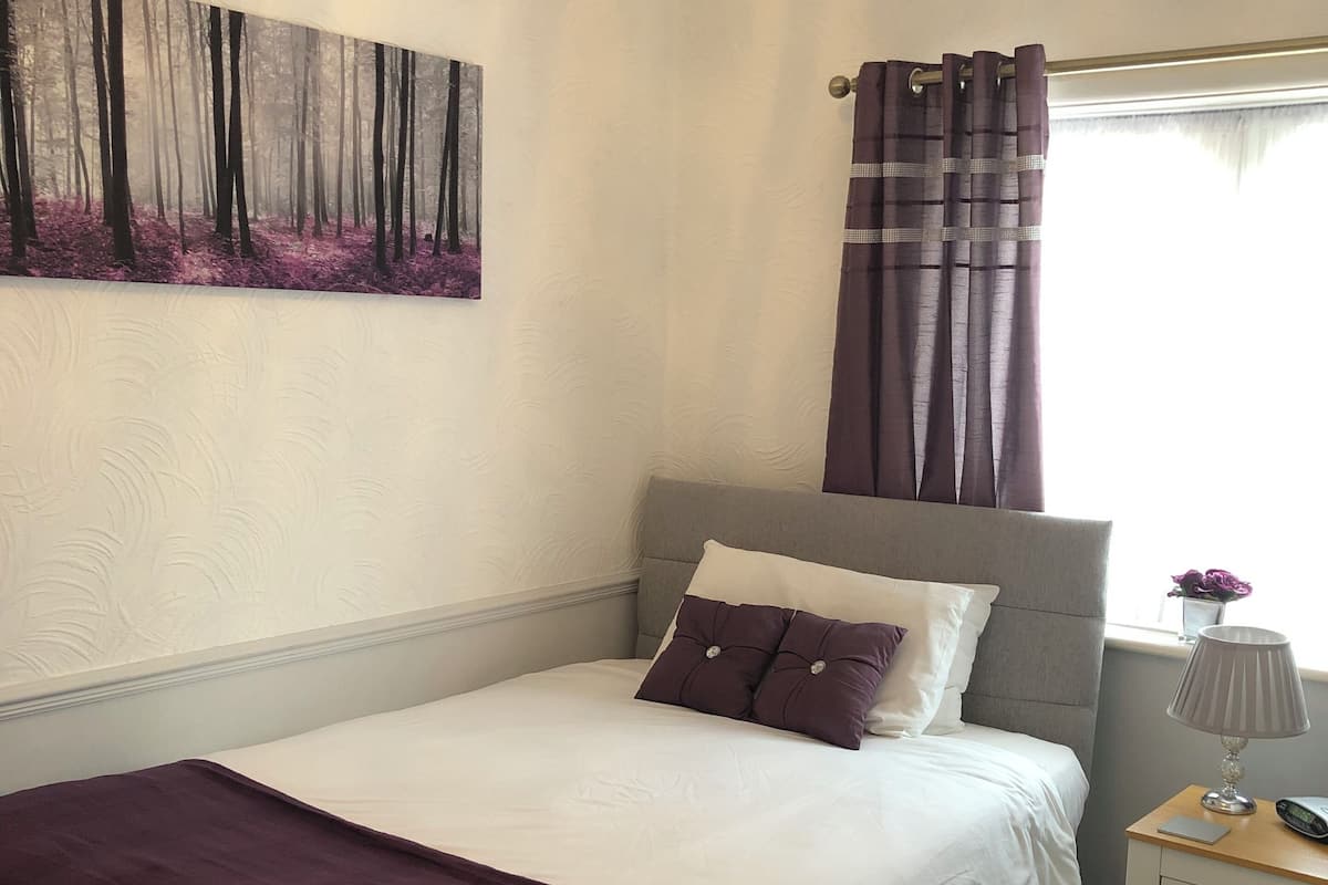 Una cama bien hecha con una colcha y almohadas moradas, una mesita de noche con una lámpara, y un cuadro de un bosque en la pared.