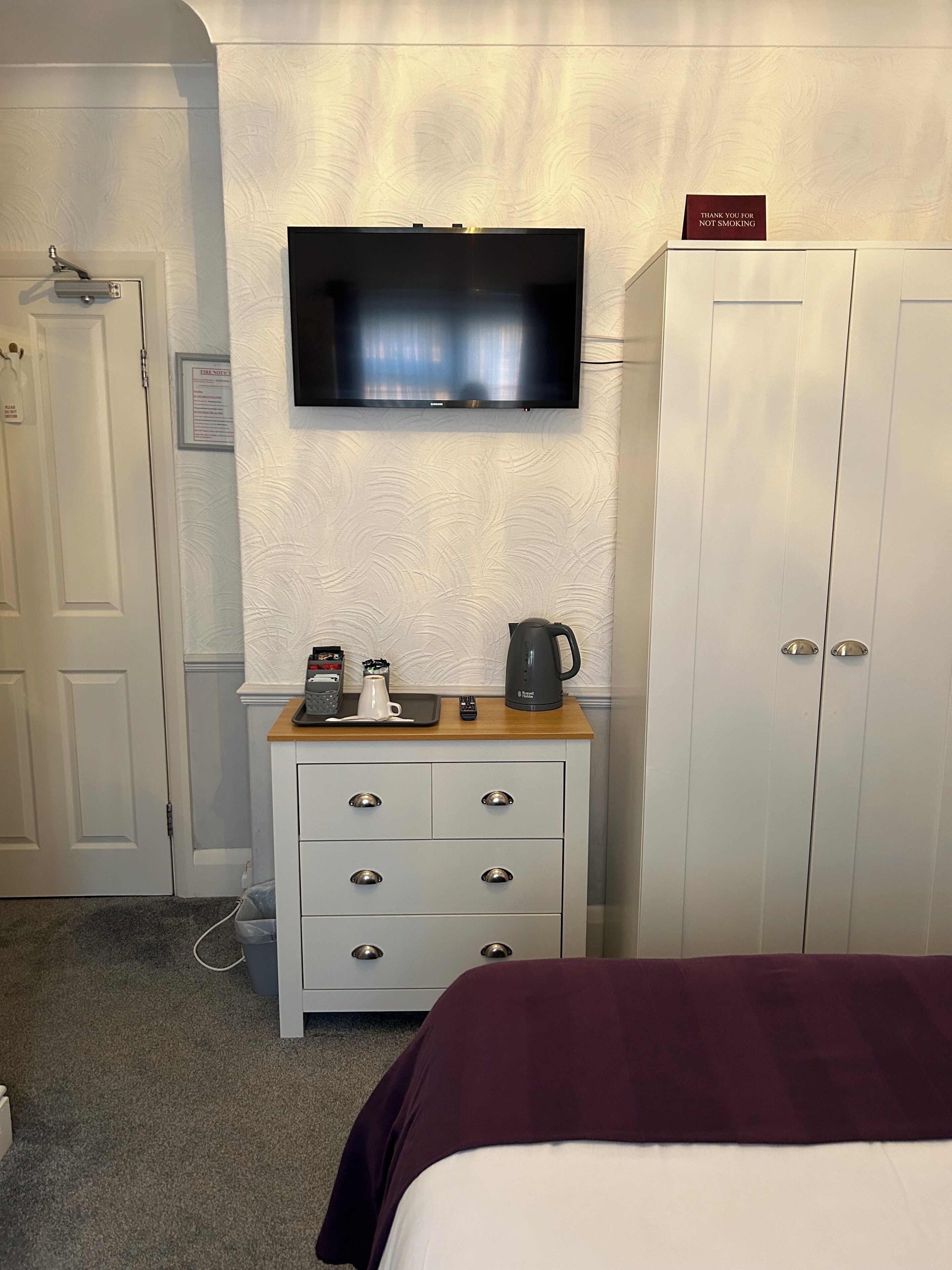 Habitación de hotel con televisión montada en la pared, minibar, cama con una manta oscura y un armario.