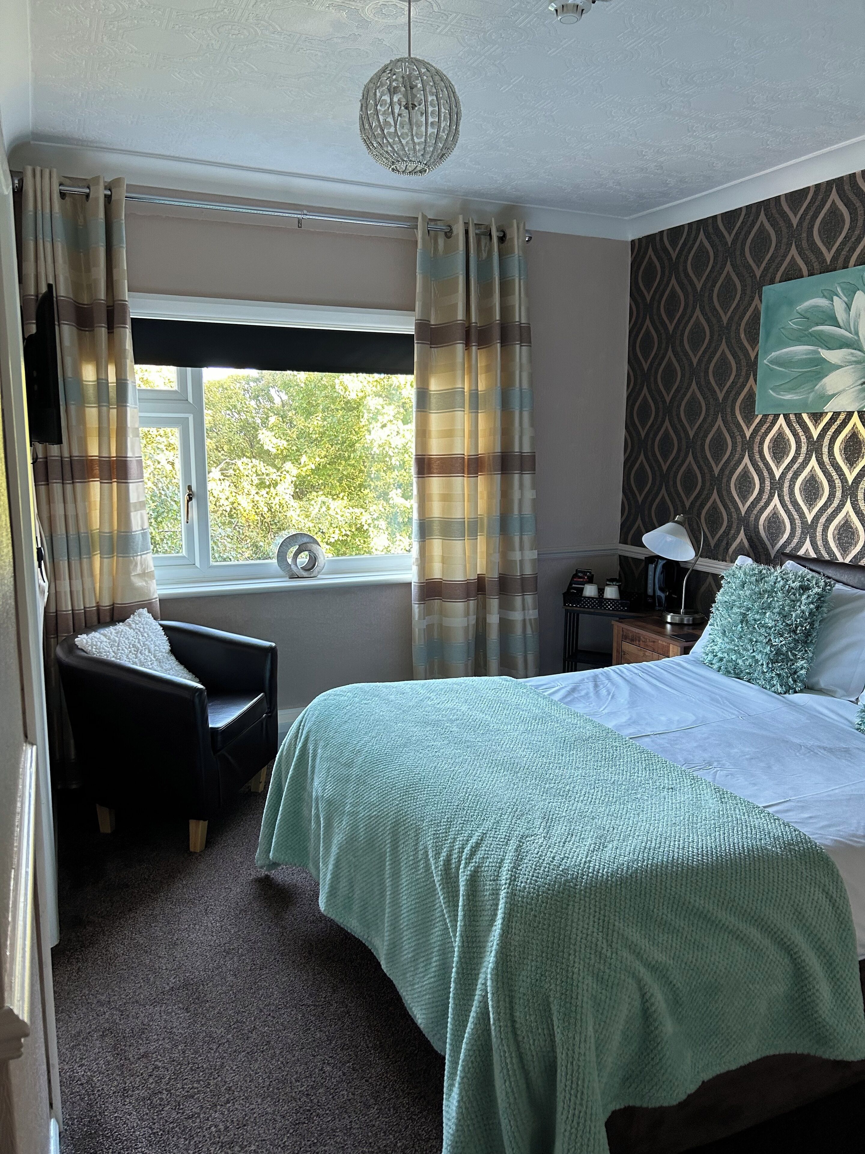 Une chambre à coucher avec un lit, une chaise, une table de chevet, une lampe et une fenêtre avec des rideaux.