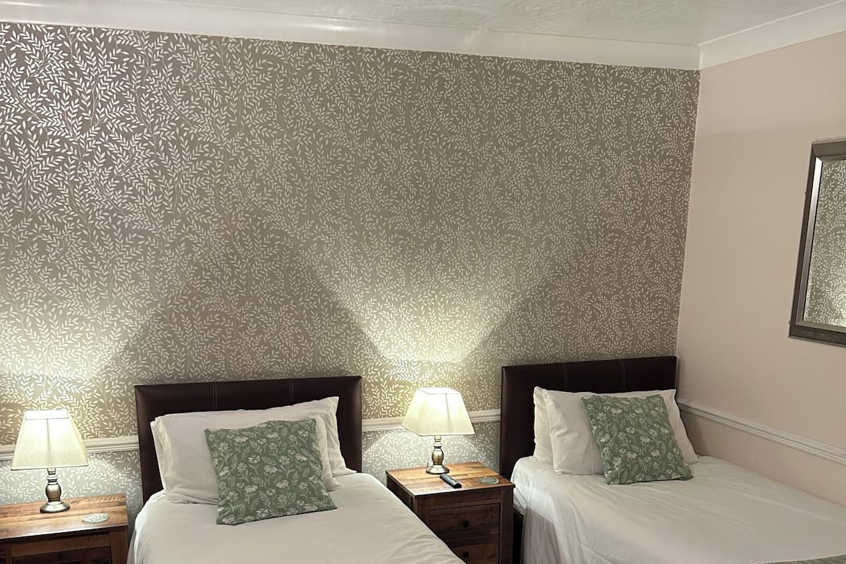Habitación de hotel con dos camas, cada una con una mesita de noche de madera y una lámpara. Las paredes tienen un papel tapiz estampado, y hay un cuadro en la pared.