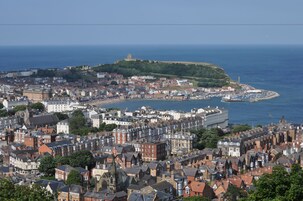 Un paisaje urbano costero con edificios históricos, un castillo destacado en una colina y un puerto deportivo con barcos.