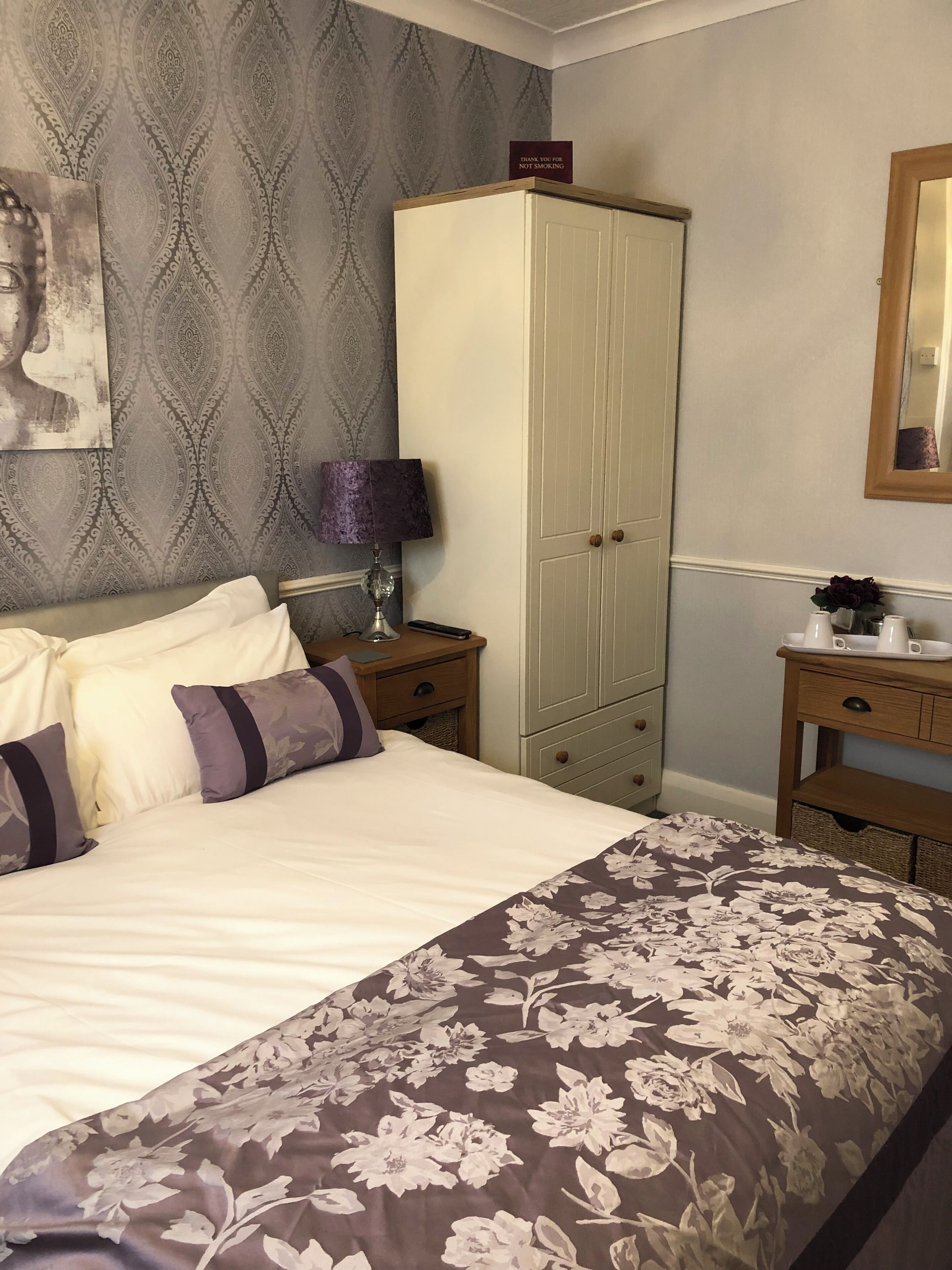 A neatly made bed with a floral duvet, a bedside lamp, a wardrobe, and a small table with a vase and cups.