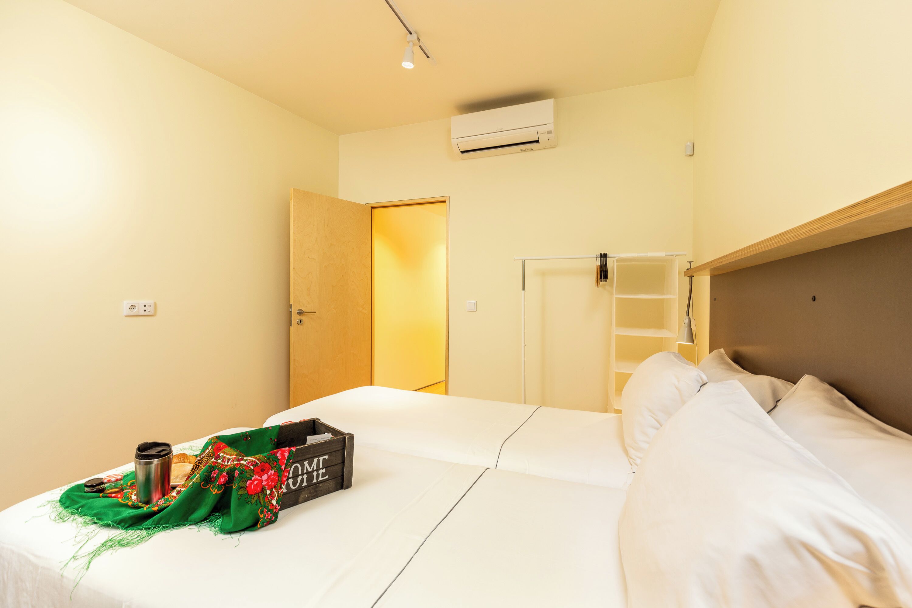 Une chambre d’hôtel avec deux lits, une tête de lit en bois, une armoire et une petite table sur laquelle se trouvent une tasse et une boîte.