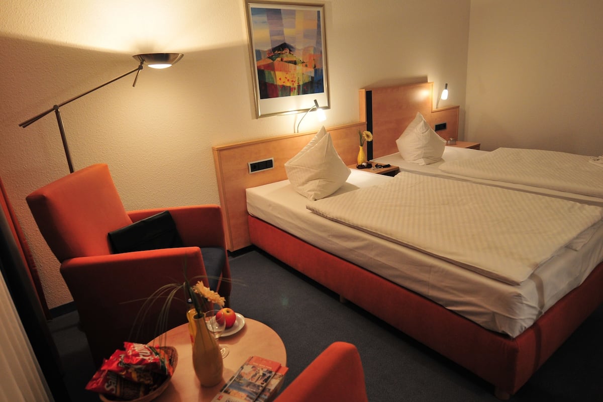Habitación de hotel con cama doble, dos almohadas, mesita de noche con lámpara, una silla y una pequeña mesa con un florero y frutero.