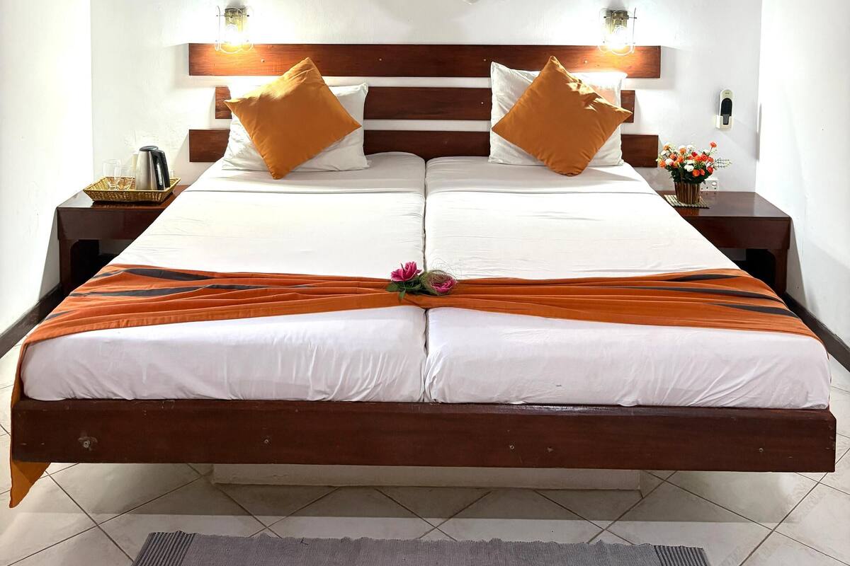 Een tweepersoonsbed met houten hoofdbord, wit beddengoed, twee oranje kussens, een decoratief middelpunt en een klamboe erboven.