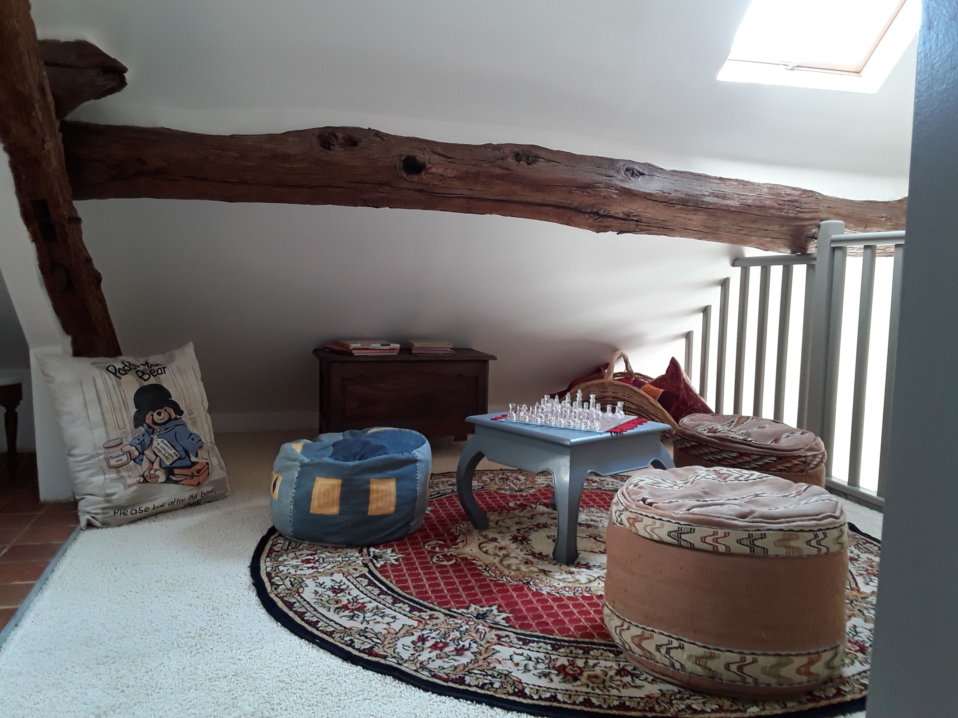 Um quarto aconchegante com teto de vigas de madeira, uma pequena mesa com peças de xadrez, um tapete estampado e uma poltrona de cadeira de saco azul.