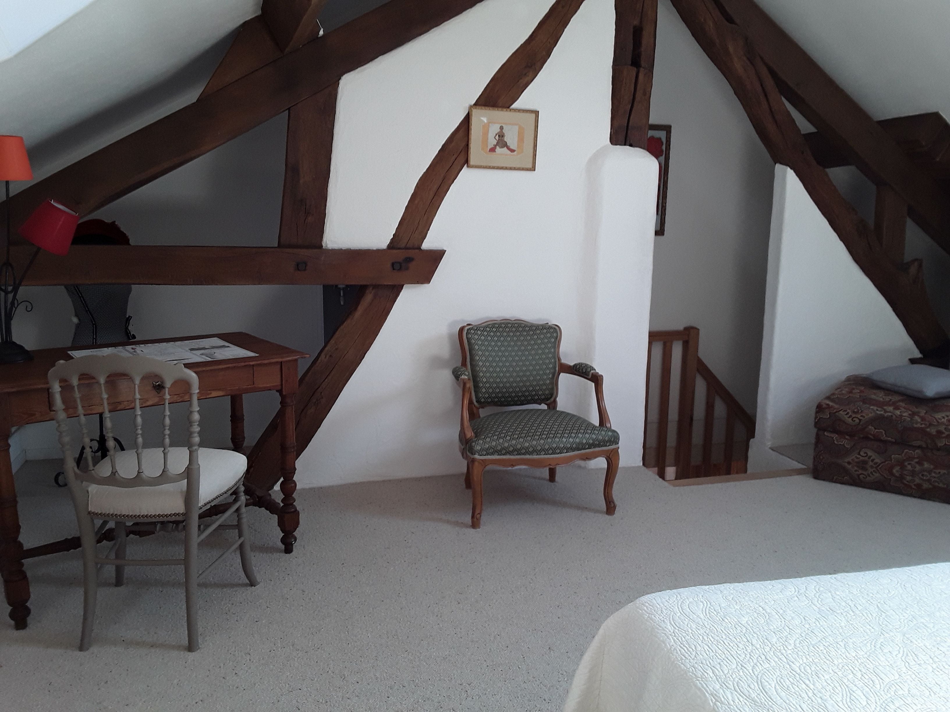 A room with a sloped ceiling, wooden beams, a desk with papers, a chair, a cushioned bench, and a framed picture on the wall.