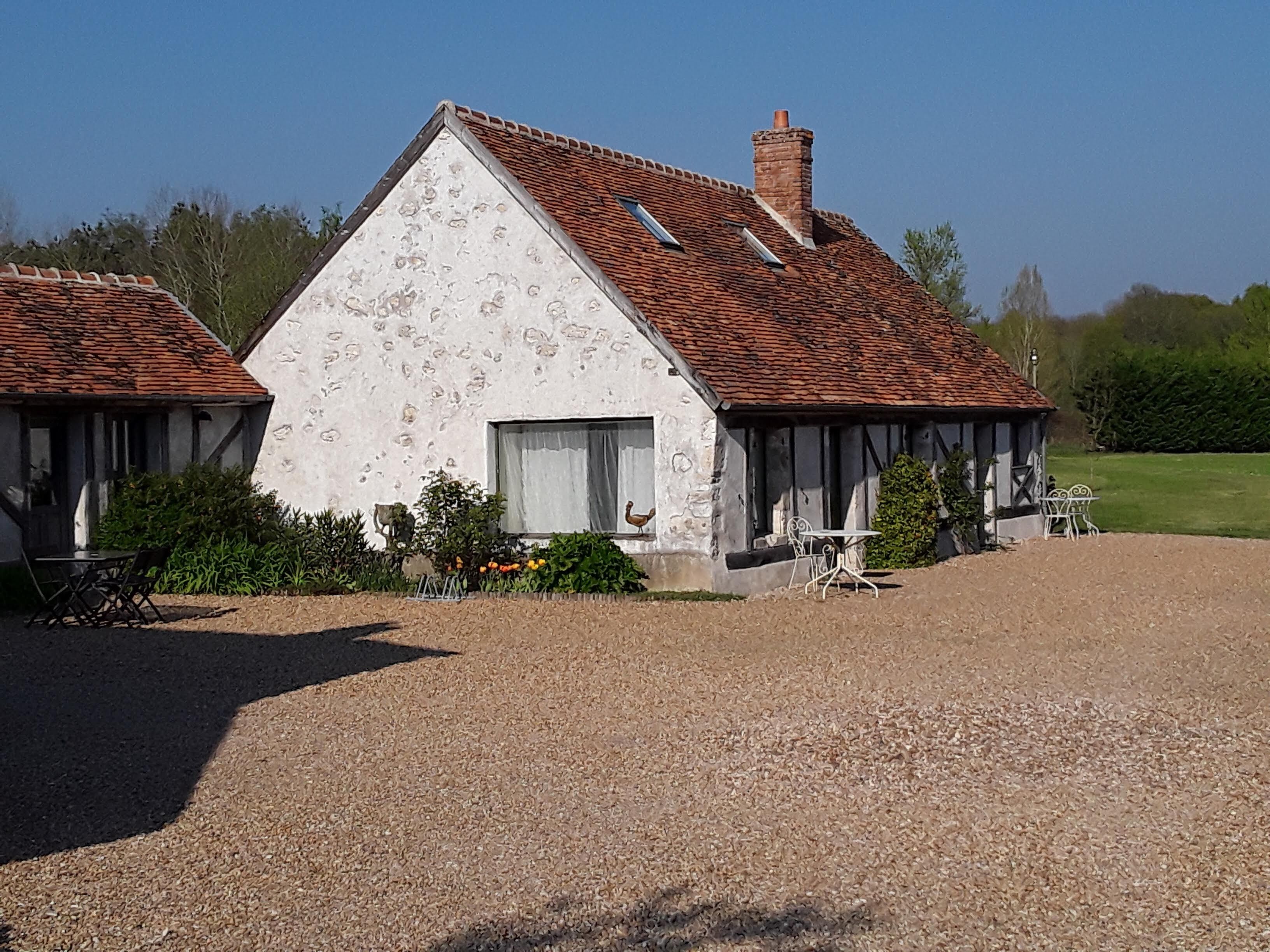 A rustic building with a sloped roof and a chimney, surrounded by a gravel driveway and a small garden area with outdoor furniture.