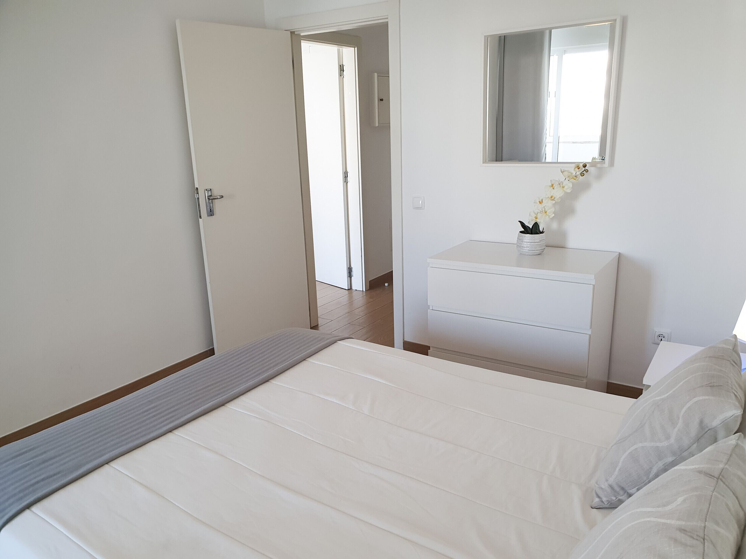 Quarto com cama, cômoda, espelho e porta que dá para outro espaço.