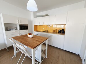 A modern kitchen with white cabinets, a wooden dining table, and a bowl of fruit.