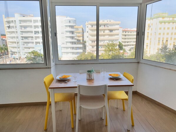 A dining area with a table and chairs, a window with a view of buildings, and a small plant on the table.