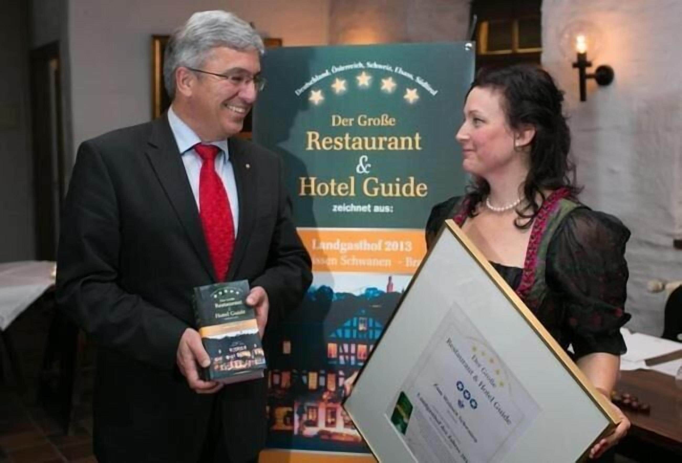 Un hombre y una mujer sostienen un documento enmarcado y un libro, de pie frente a un letrero que dice "Der Große Restaurant & Hotel Guide".