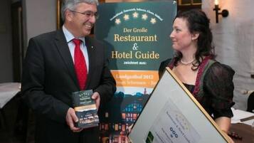 Un hombre y una mujer sostienen un documento enmarcado y un libro, de pie frente a un letrero que dice "Der Große Restaurant & Hotel Guide".