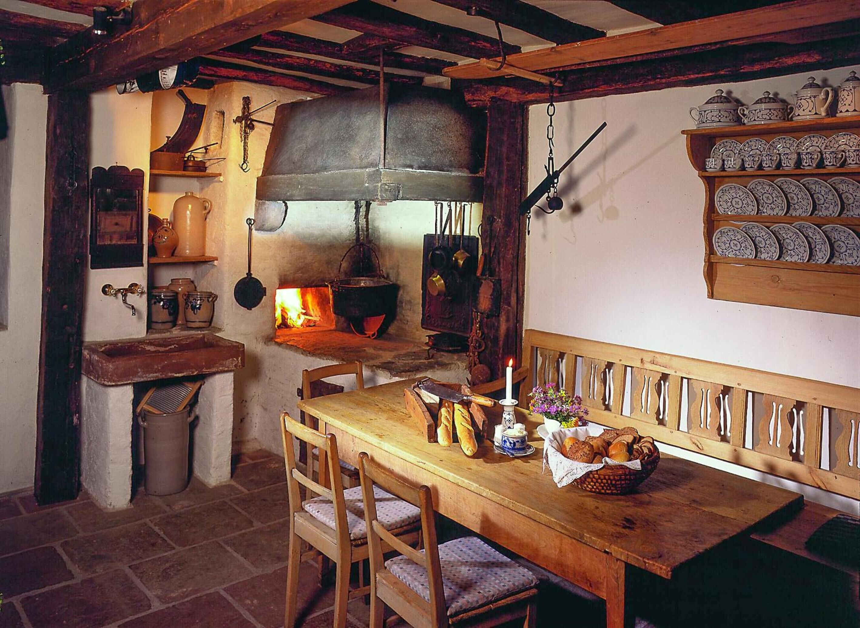 A rustic kitchen with a wooden table, chairs, a hanging pot rack, and a shelf with ceramic dishes.
