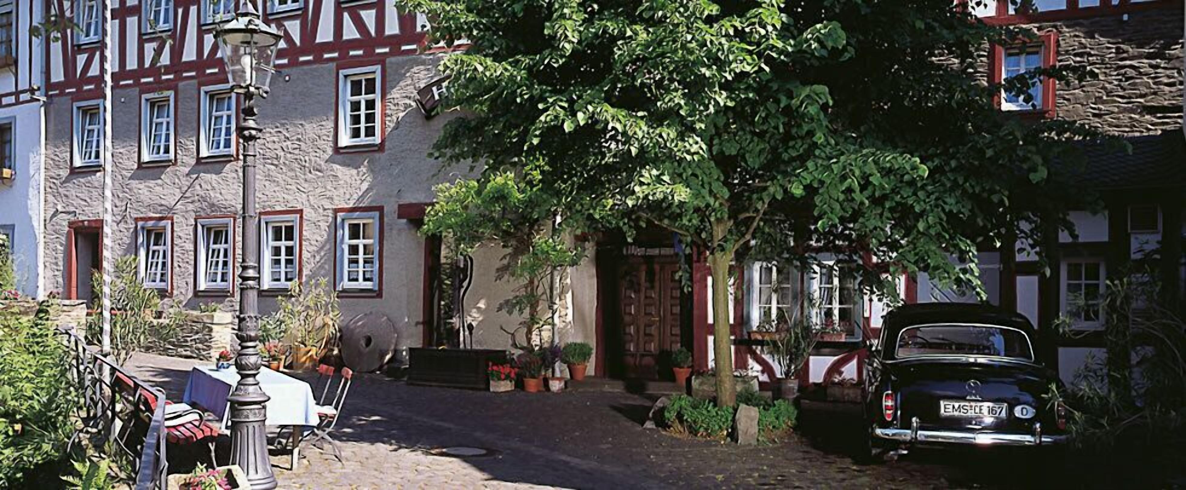 A cobblestone street with a vintage car parked on the right. There are buildings with traditional architecture, including a house with a stone facade and a window with a red frame.
