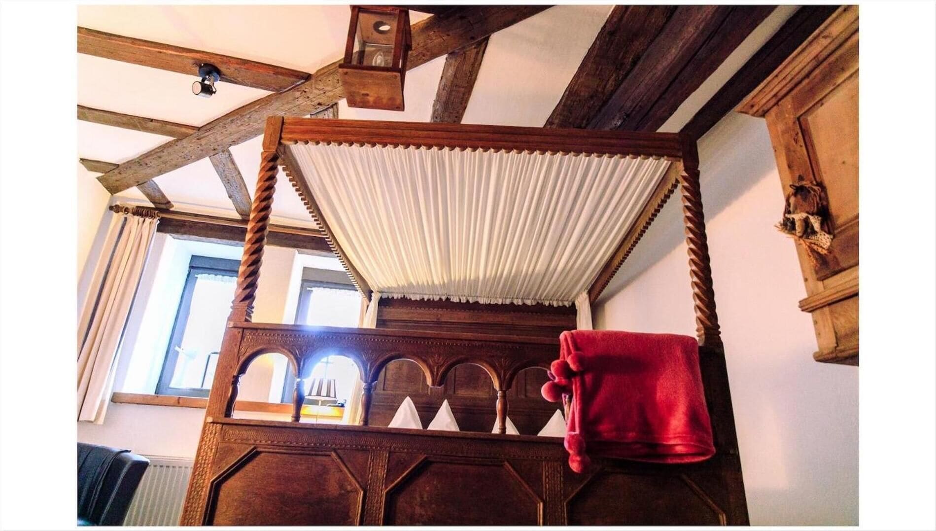 Uma cama de madeira tradicional com dossel, uma toalha vermelha e teto rústico com vigas aparentes.