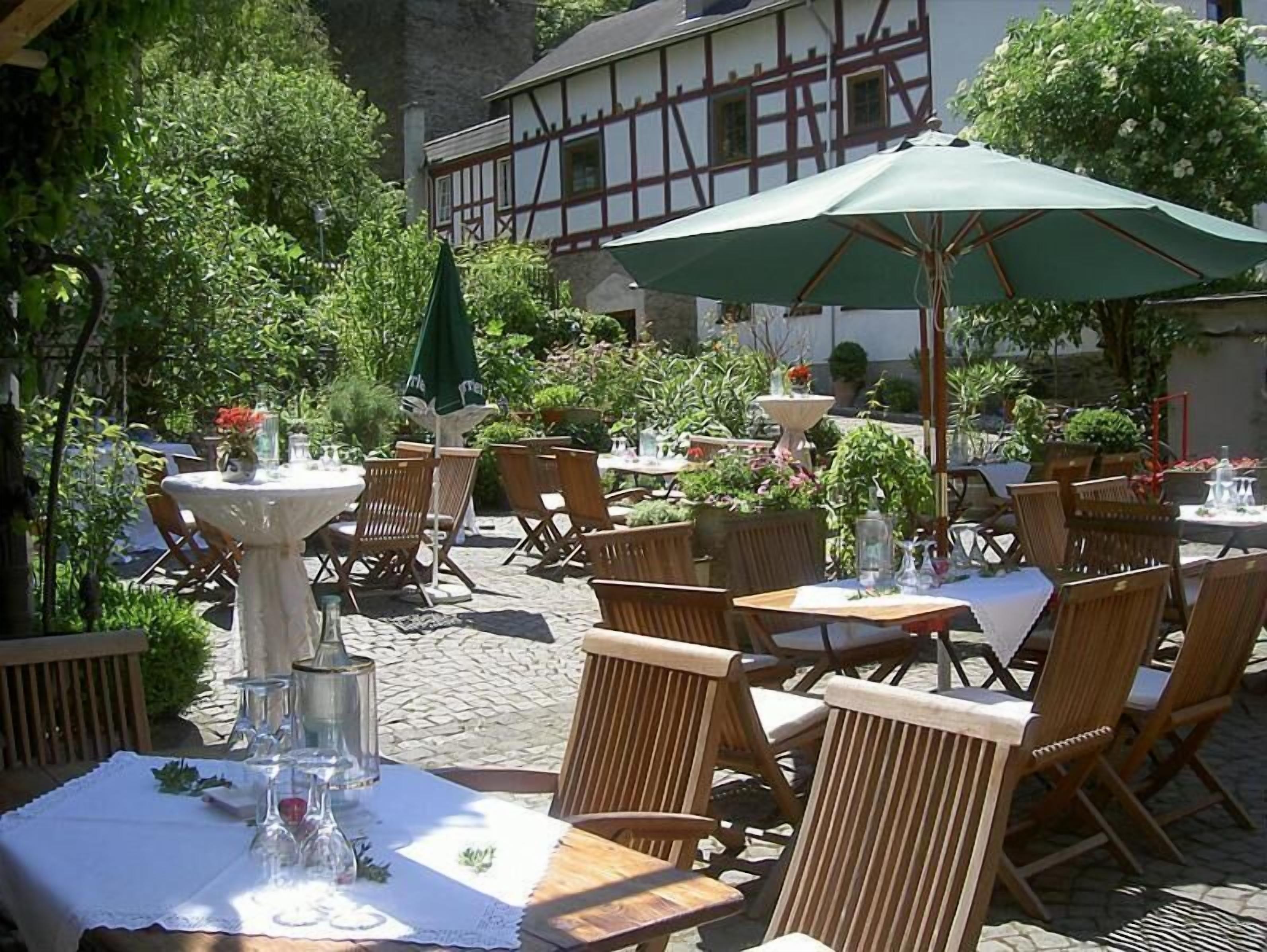Ein Essbereich im Freien mit Tischen und Stühlen, umgeben von viel Grün und einem Gebäude mit traditioneller Fassade.