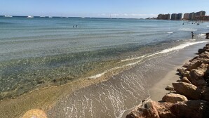 Una playa con agua cristalina, costa rocosa y edificios a lo lejos.