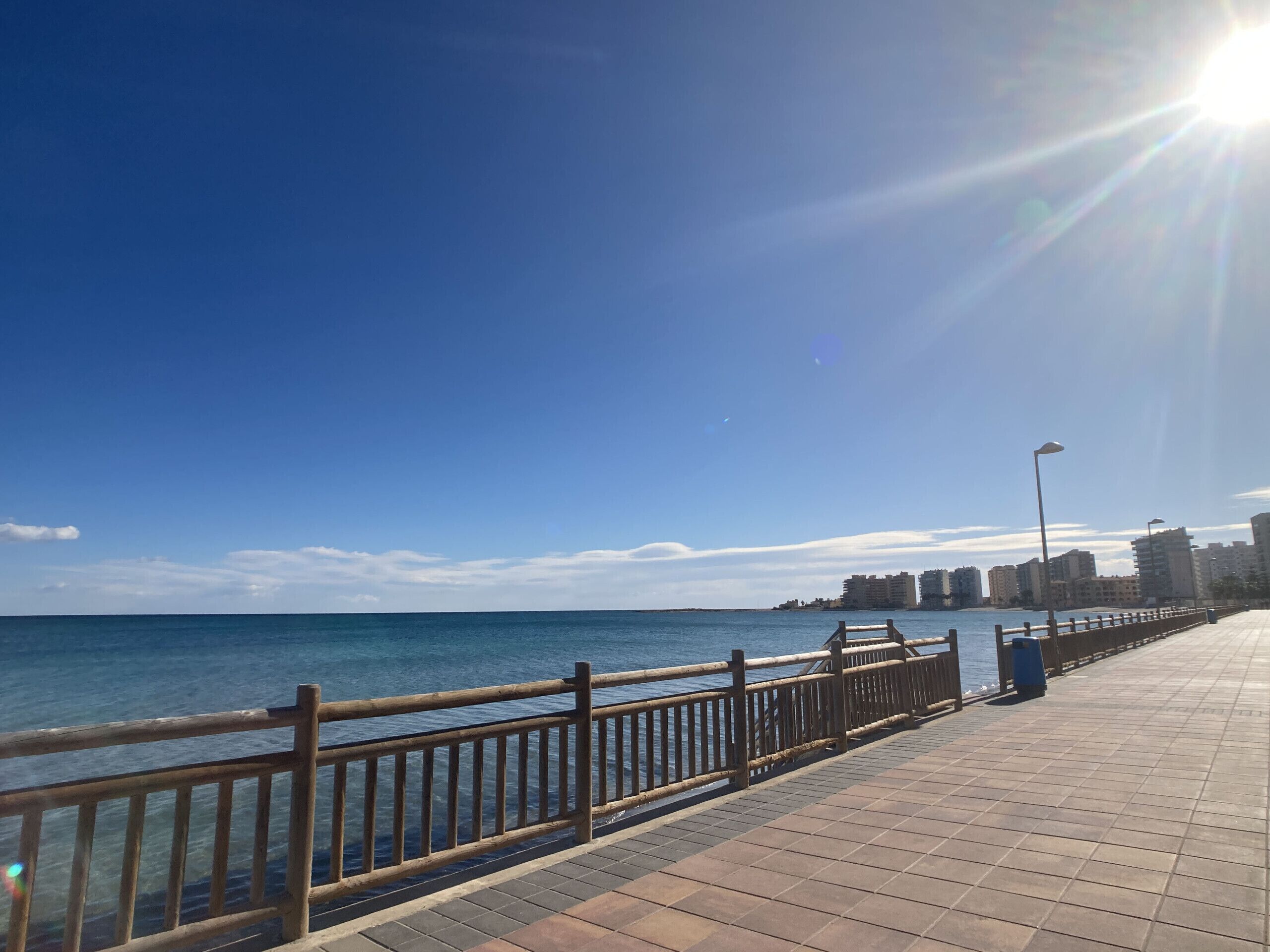 A coastal promenade with a wooden railing, a tiled walkway, and a clear view of the sea and distant buildings.