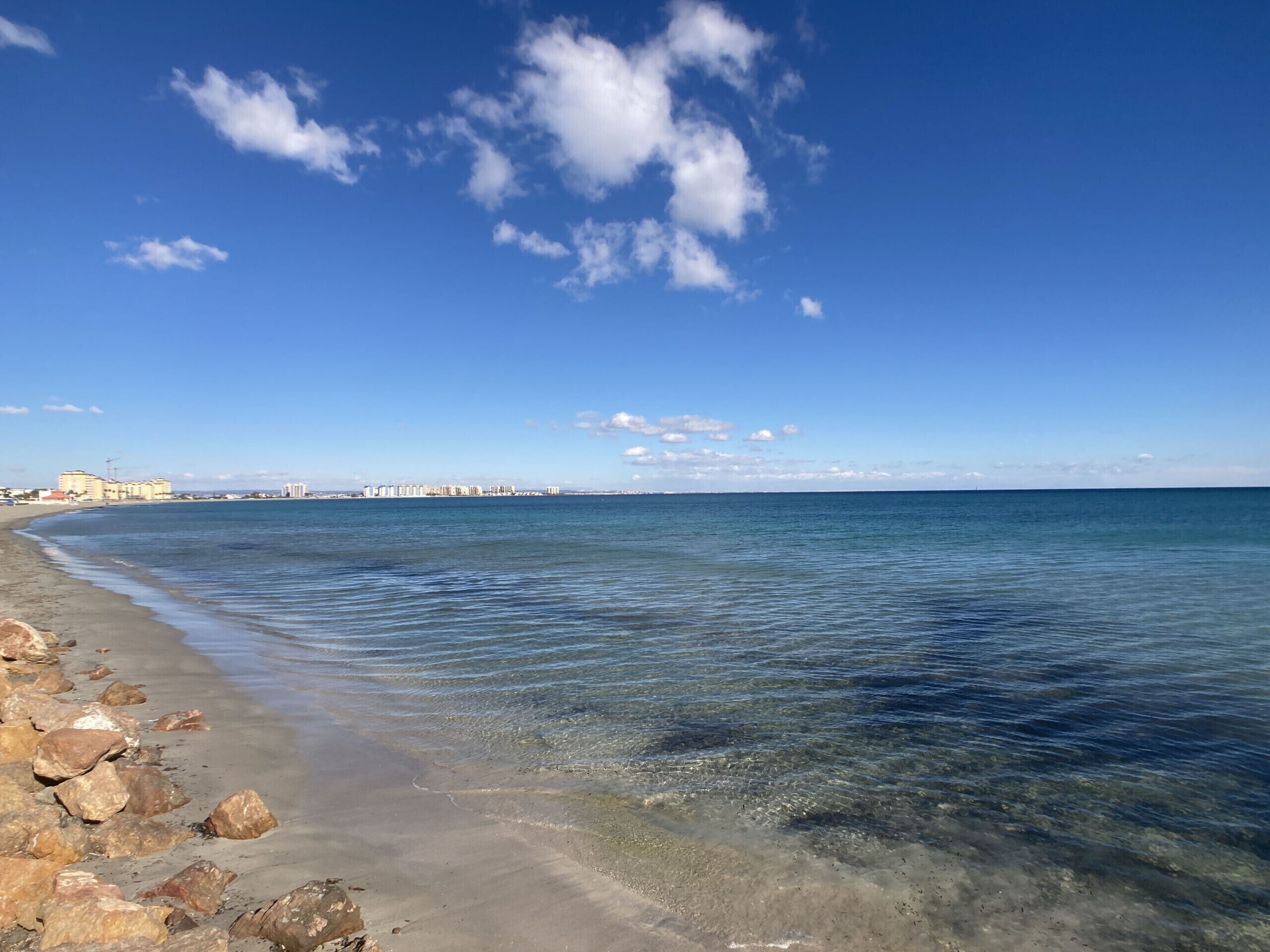 A beach with clear water, a rocky shoreline, and buildings in the distance.