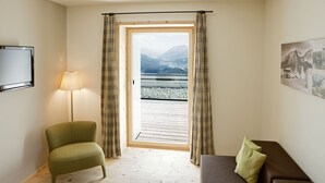 A room with a view of mountains and a lake through a window, a green chair, a brown sofa with a pillow, and a lamp.