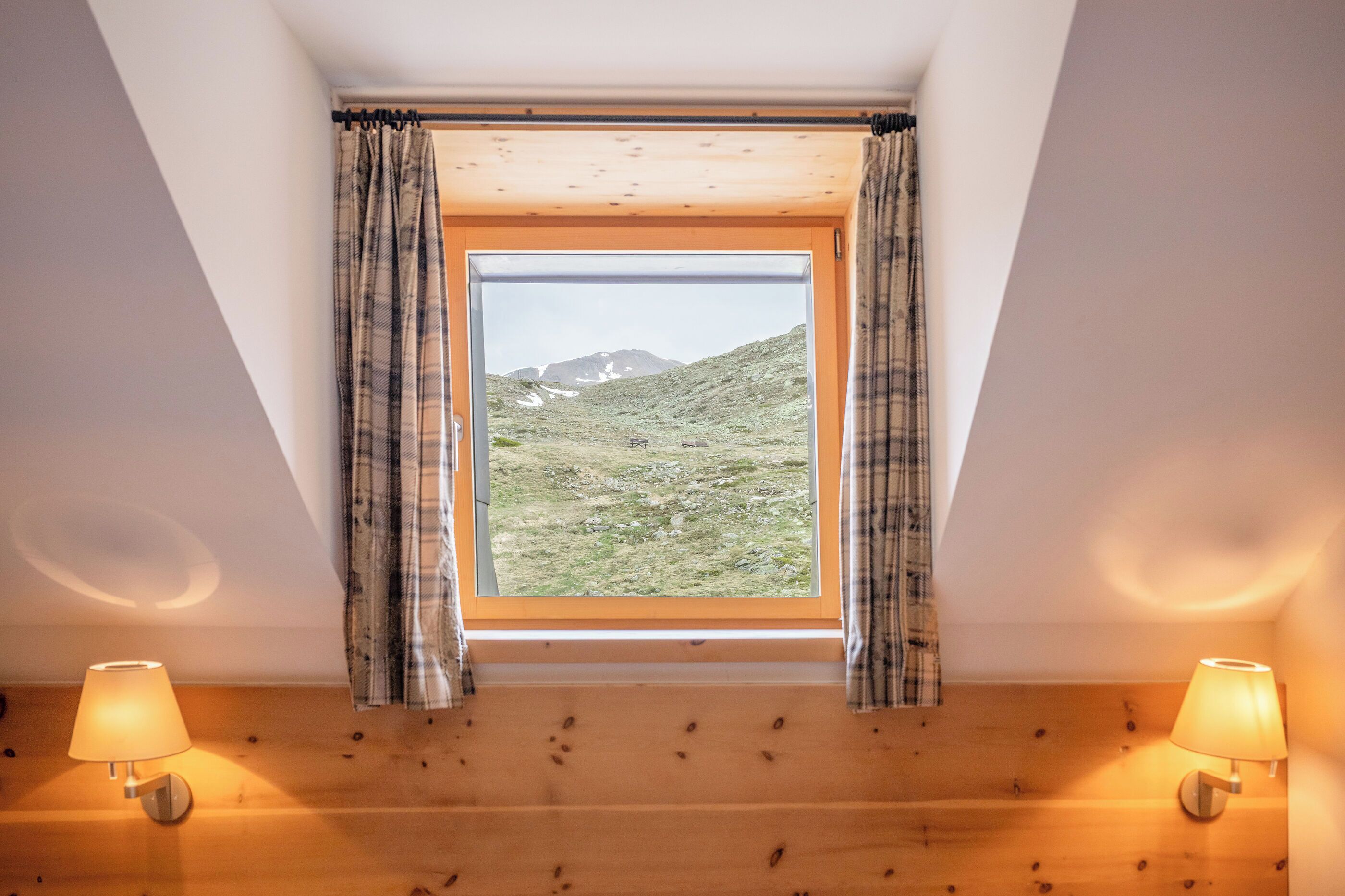 A room with a wooden sloped ceiling, a window with plaid curtains, and two wall-mounted lamps.