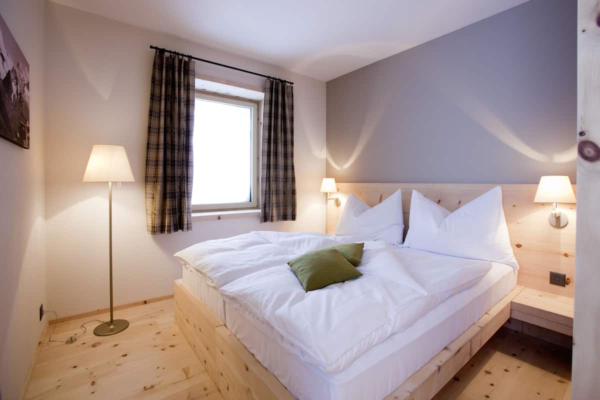 Un dormitorio con piso de madera, una cama con ropa blanca y un almohón verde, dos lámparas de noche, una ventana con cortinas escocesas y una lámpara de pared.