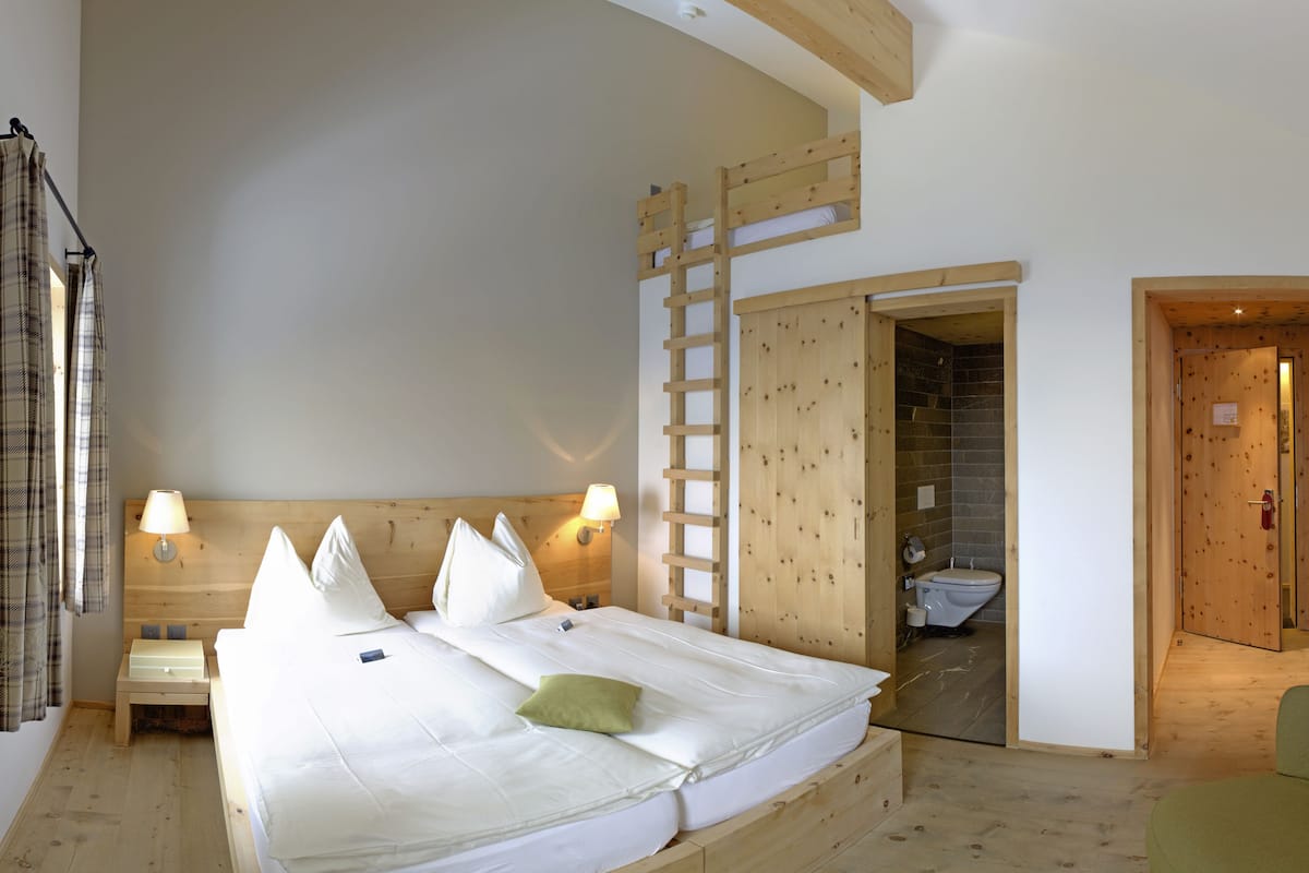 Una habitación de madera con una cama, dos lámparas de noche, una escalera de madera y un baño visible a través de una puerta abierta.