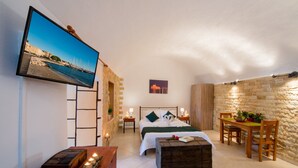 A cozy bedroom with a bed, bedside tables, a TV, a wooden chest, and a stone wall. - Emerald Dream House (Rhodes)