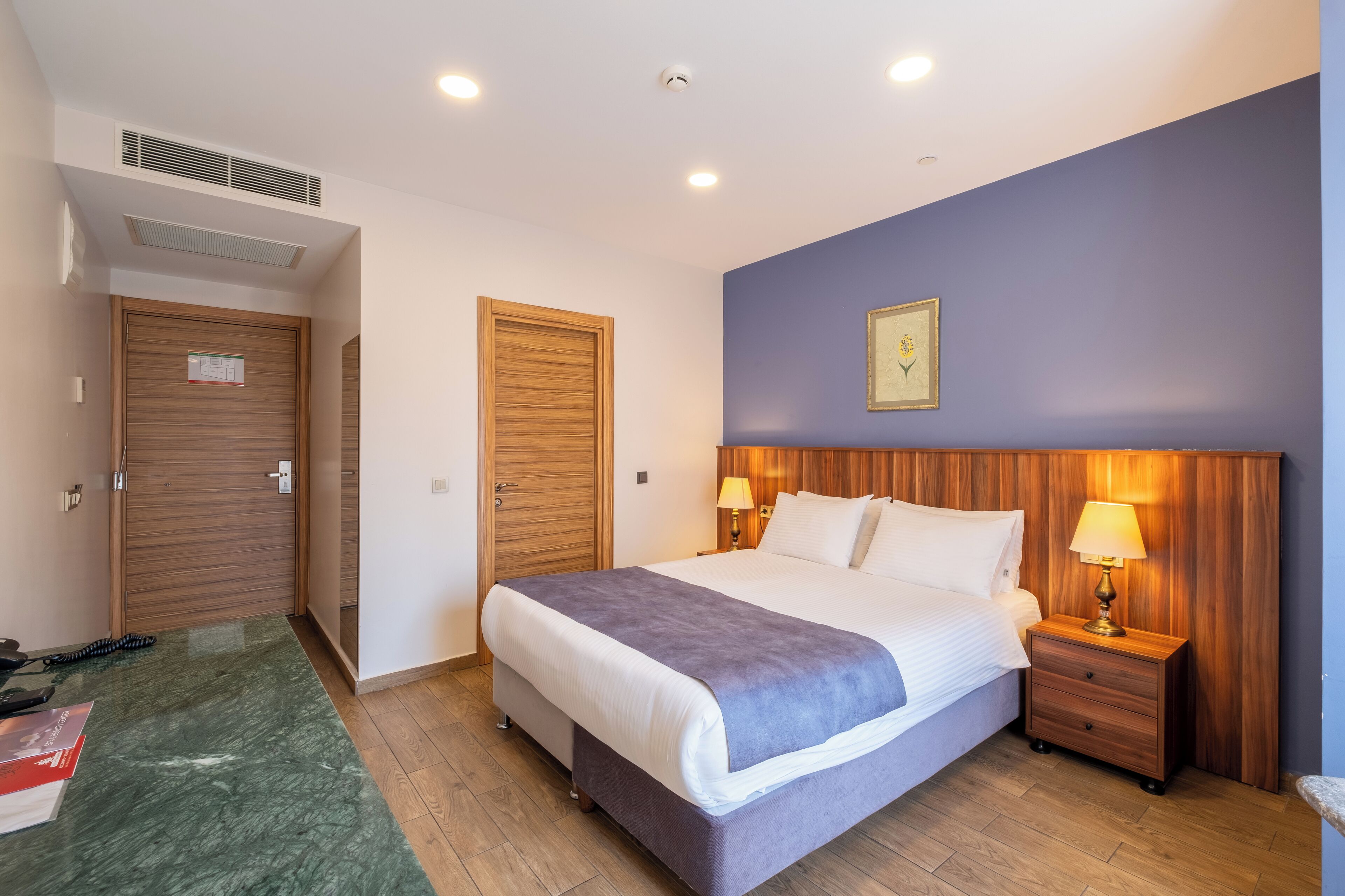 Habitación de hotel con cama, cabecera de madera, dos mesitas de noche con lámparas, alfombra verde y suelo de madera.