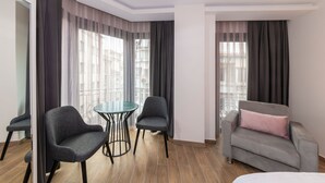 Una habitación de hotel moderna con una mesa redonda de cristal, dos sillones grises, un sofá gris con un cojín rosa y ventanas amplias con cortinas de gasa.