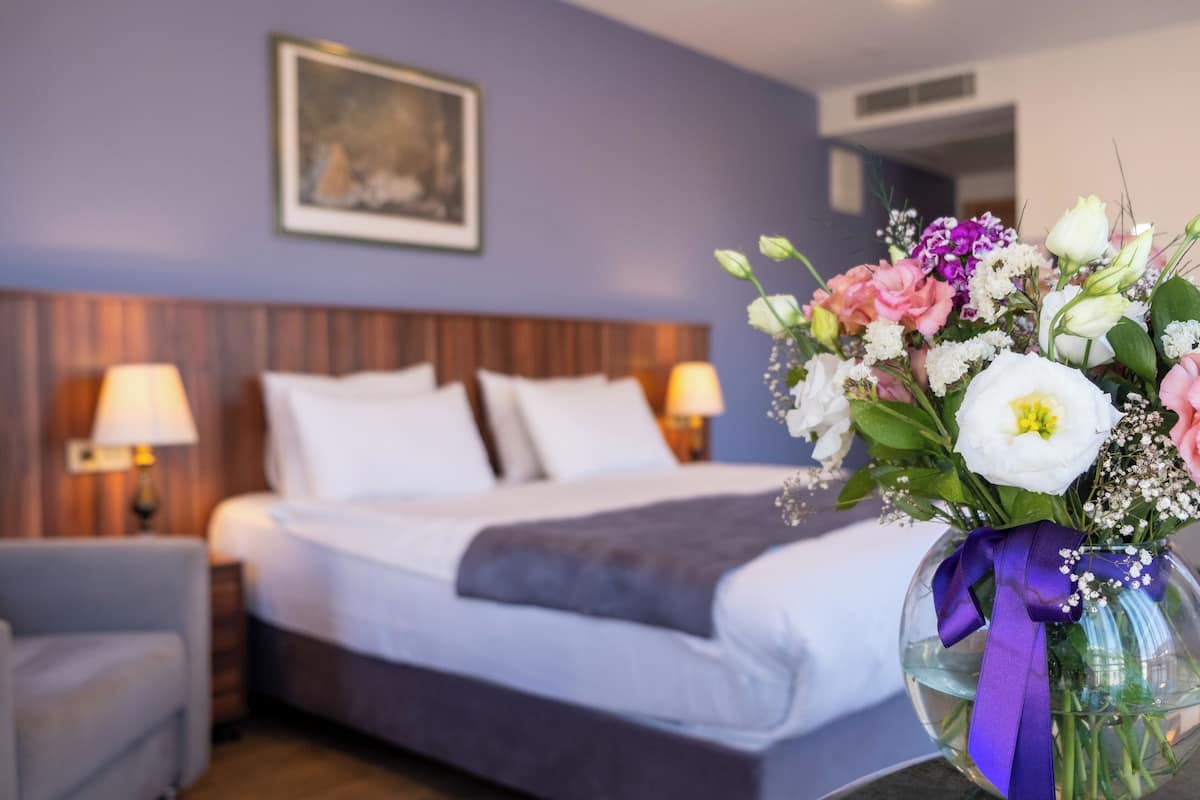 A hotel room with a bed, two bedside lamps, a framed picture on the wall, and a vase with flowers on a table.