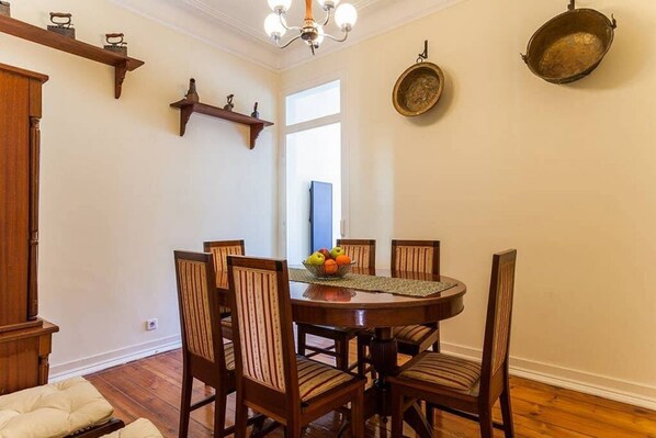 A dining area with a round wooden table, six chairs, and a fruit bowl on the table. There are two wall-mounted shelves above the table, and a large window with a view of the outside.