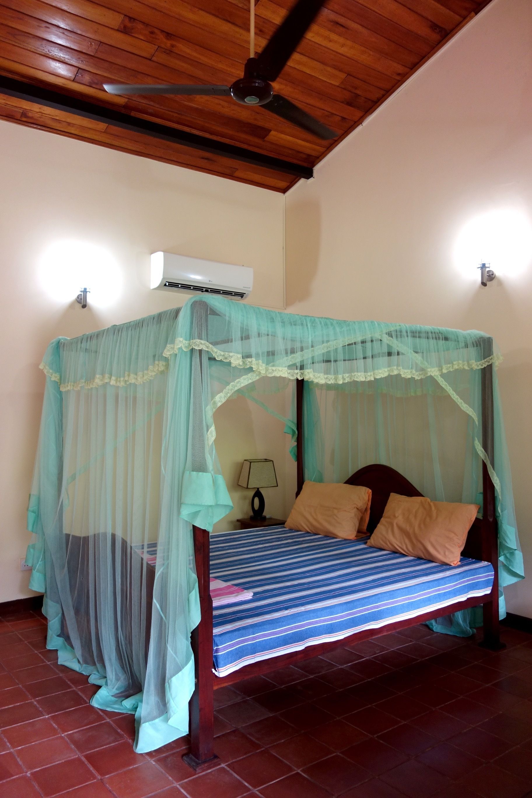 A bed with a canopy, a ceiling fan, an air conditioner, and a lamp on the wall.
