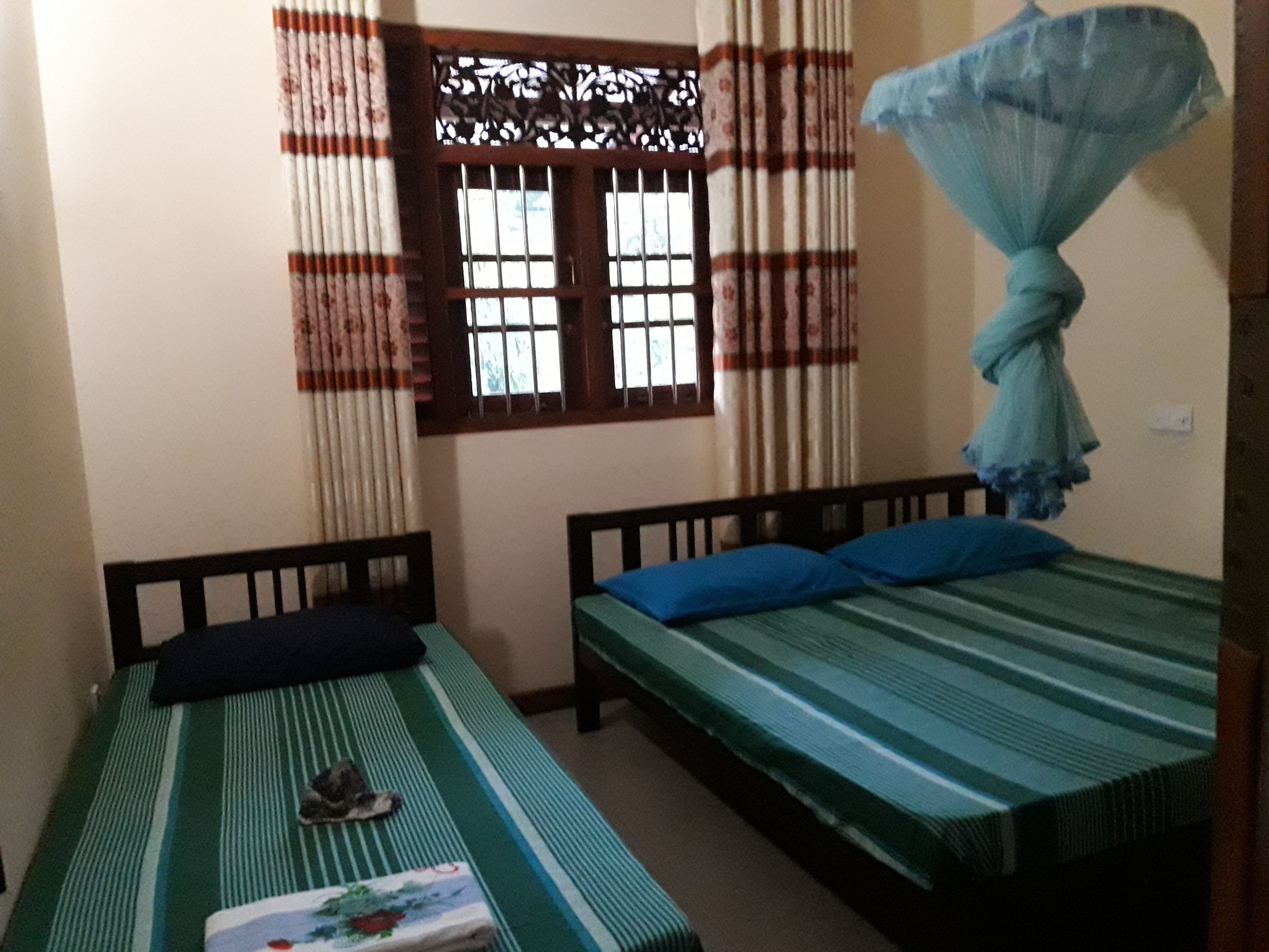 A room with two single beds, one with a striped bedspread and the other with a solid green bedspread. There is a window with a decorative grille and curtains, and a mosquito net hanging on the wall.