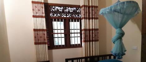 A room with two single beds, one with a striped bedspread and the other with a solid green bedspread. There is a window with a decorative grille and curtains, and a mosquito net hanging on the wall.