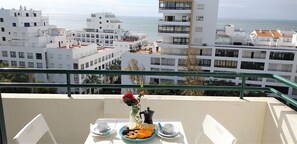 A balcony with a table set for breakfast, overlooking a coastal cityscape with modern buildings and the sea.