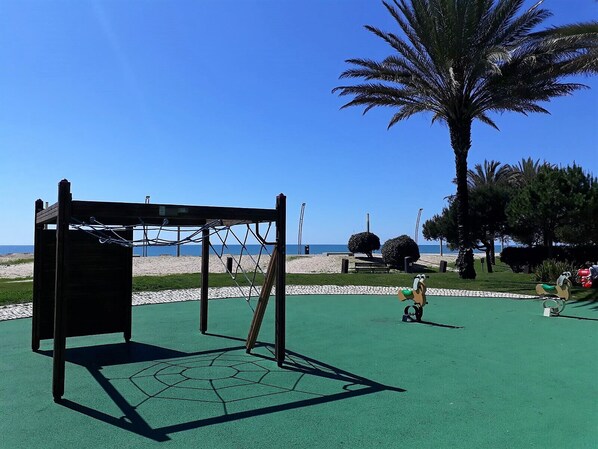 A playground with a climbing structure and a basketball hoop, set against a beach backdrop.