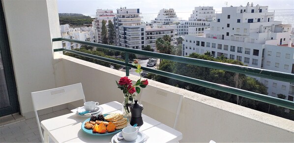 A balcony with a table set for breakfast, a vase with flowers, and a view of white buildings and the sea.