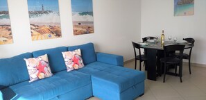 A blue sofa with starfish pillows, a dining table with chairs, and three beach-themed paintings on the wall.